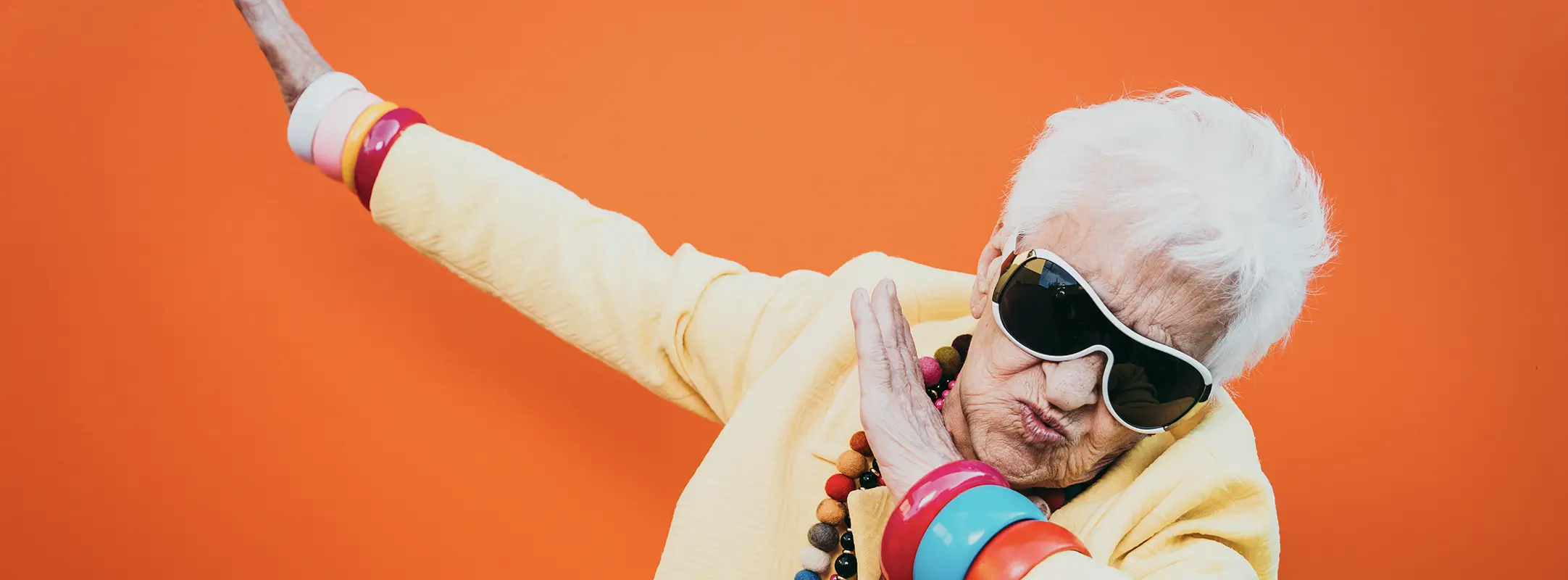 Oudere vrouw met haar handen in de lucht met zonnebril op en kettingen om.