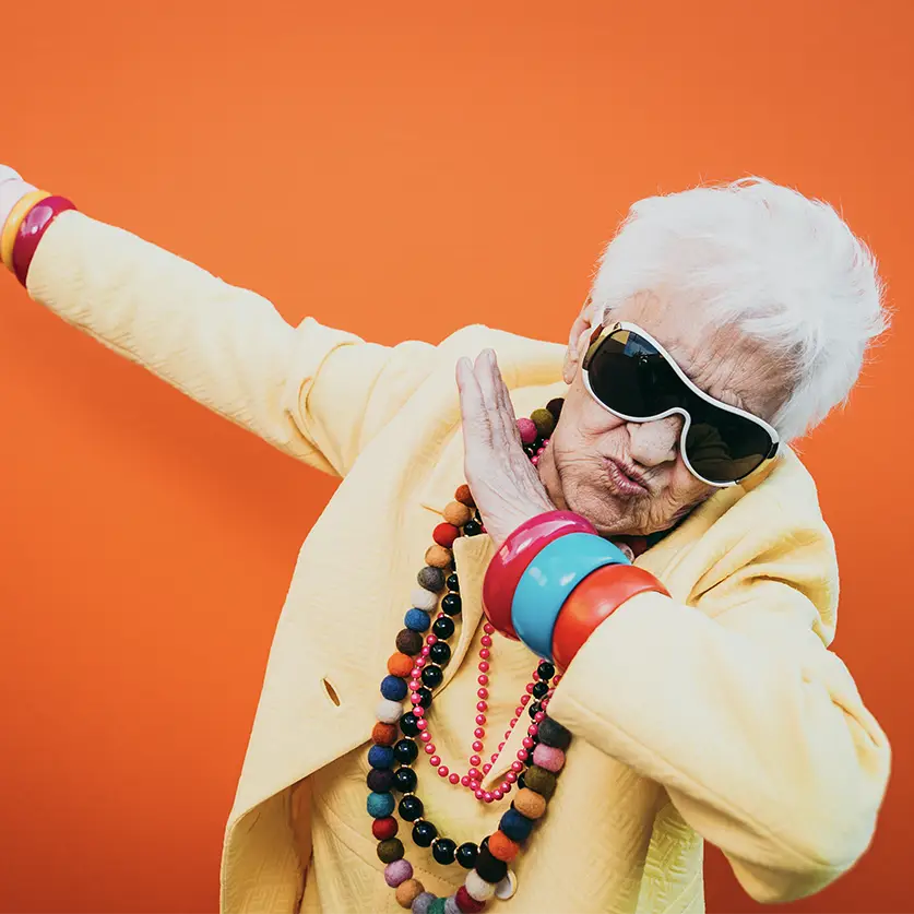 Oudere vrouw met haar handen in de lucht met zonnebril op en kettingen om.